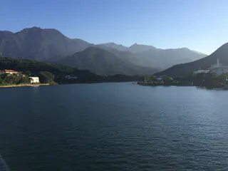 A calm lake with gentle ripples, surrounded by green hills and distant mountains under a clear blue sky. Buildings and trees line the shore on the left and right sides. The sunlight casts long shadows over the landscape.