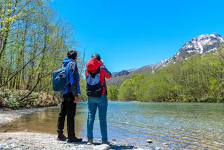 Two people in jackets and backpacks stand by a clear river, looking at a snow-capped mountain under a bright blue sky, surrounded by green trees.