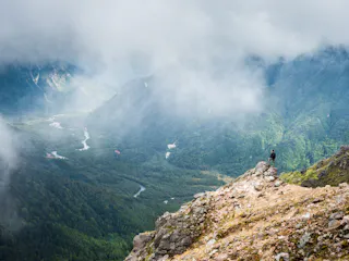 A person stands on a rocky ledge overlooking a lush green valley with winding rivers, surrounded by misty mountains and partially covered by clouds.