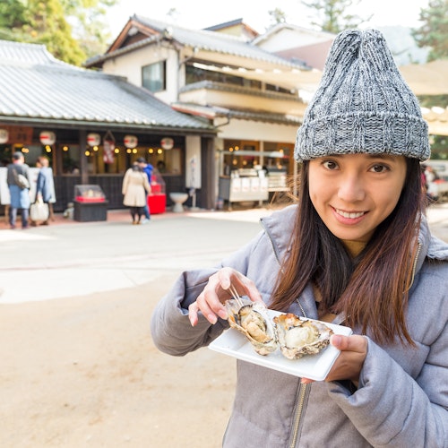 Miyajima Oyster Festival A woman in a gray coat and knitted hat smiles while holding a plate of oysters with a fork. She stands in an outdoor market area with traditional buildings and people in the background. The scene suggests a chilly day.