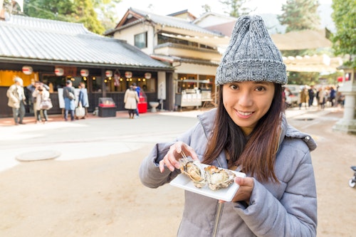 A woman in a gray coat and knitted hat smiles while holding a plate of oysters with a fork. She stands in an outdoor market area with traditional buildings and people in the background. The scene suggests a chilly day.