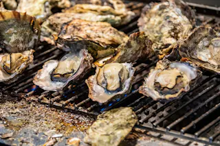 Open oysters are being grilled on a barbecue, with some shells open to reveal the cooked oysters inside. The grill has visible flames and bits of shell scattered around.