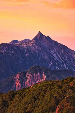 A rugged mountain peak rises dramatically under a vivid orange and pink sunset sky, with lush green hills in the foreground and rocky slopes in the background.