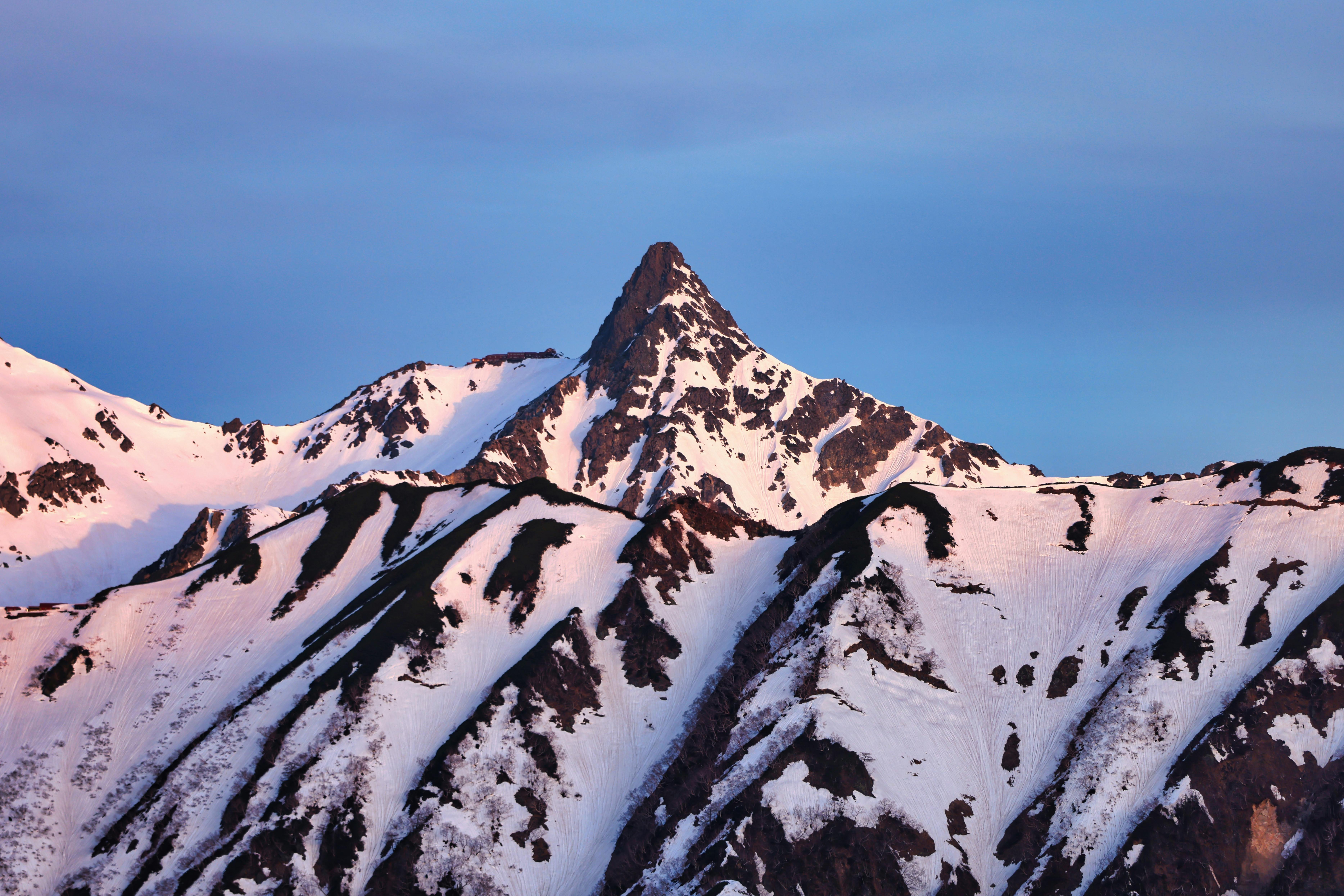 A snow-covered mountain peak is illuminated by soft pink and blue light, with rugged slopes and dramatic ridges under a clear sky.