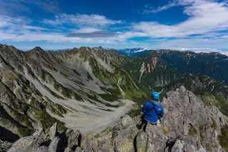 A person in a blue jacket and helmet sits on rocky terrain, overlooking a dramatic mountain landscape with jagged peaks, green valleys, and a partly cloudy blue sky.