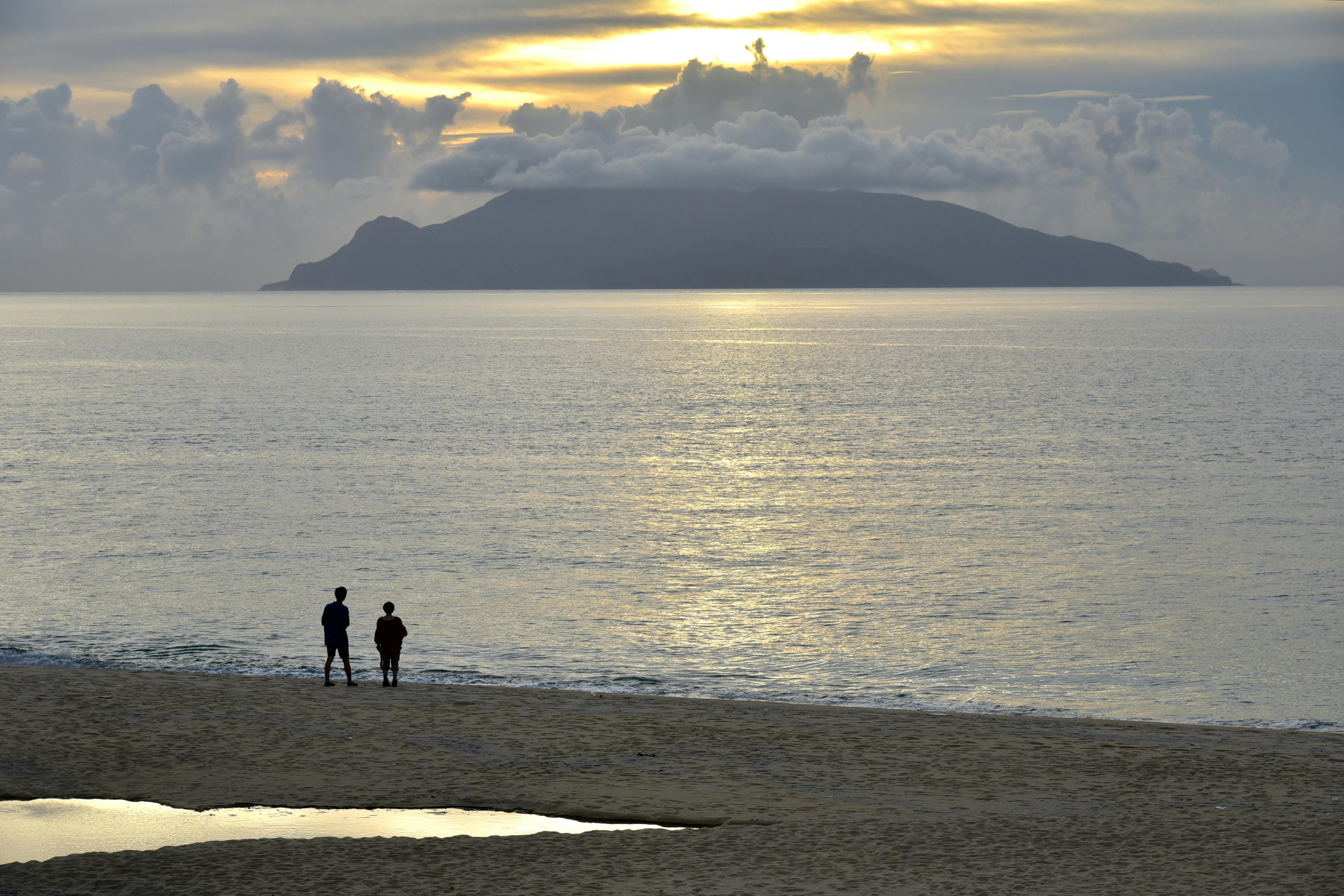 Two people stand on a sandy beach at sunset, looking out over calm water toward a distant island and cloudy sky with the sun partially hidden behind clouds.