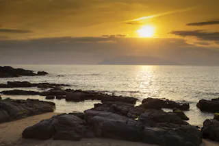Rocky shoreline at sunset, with the sun low in the sky casting golden reflections on the calm sea. Distant landmass is visible on the horizon beneath a partly cloudy sky.