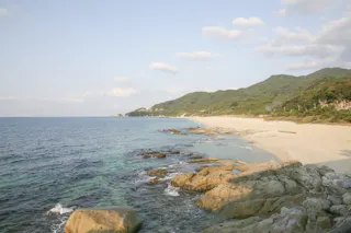 A scenic coastal landscape featuring clear blue water, rocky shore in the foreground, sandy beach, and lush green hills under a partly cloudy sky.