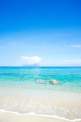 Clear turquoise sea waves gently wash onto a sandy beach under a bright blue sky. A few rocks are visible near the shore, and a distant cloud hovers above the horizon. The scene is calm and serene.