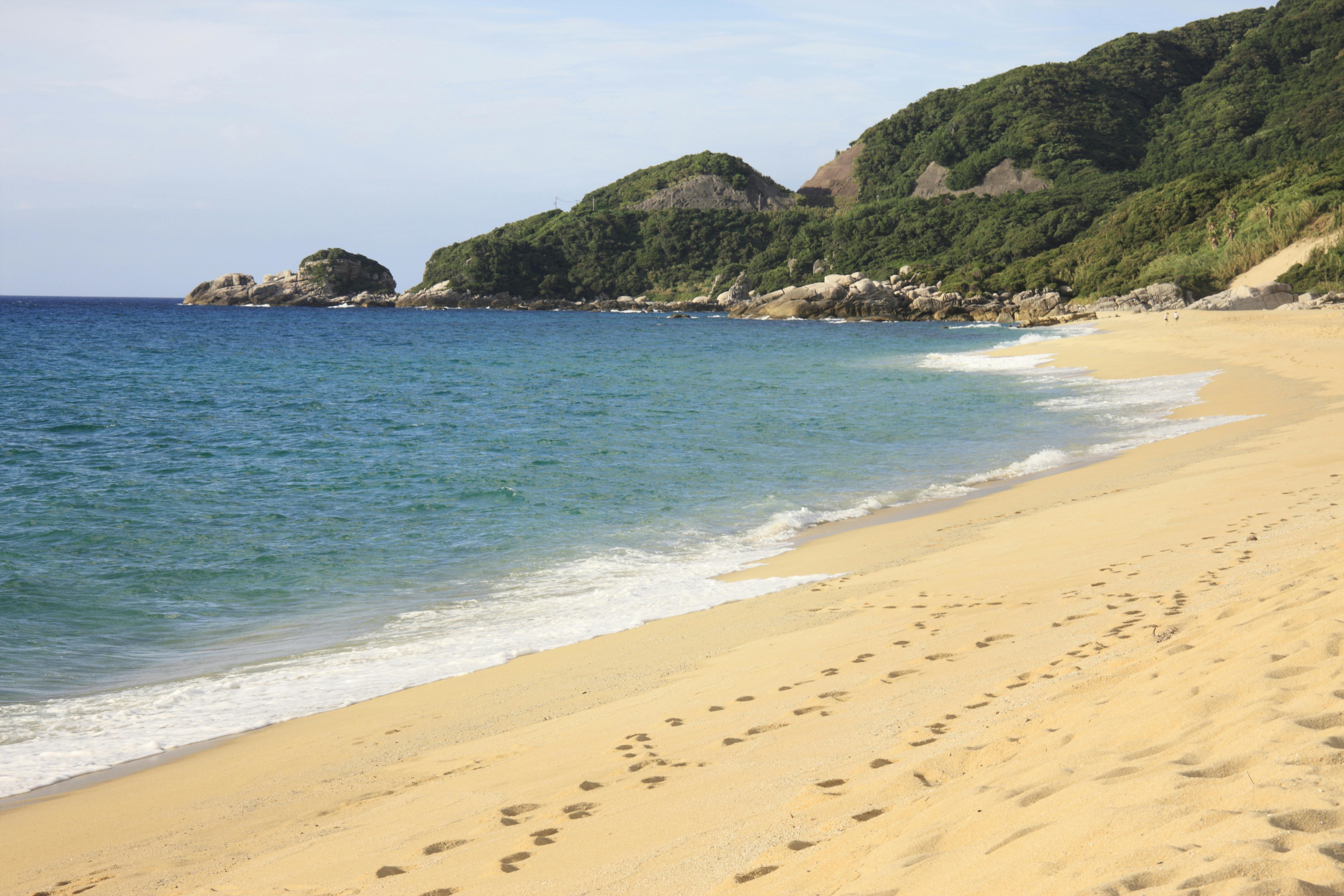 A sandy beach with gentle ocean waves, footprints leading along the shore, and green hills covered with trees in the background under a partly cloudy sky.
