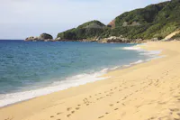 A sandy beach with gentle ocean waves, footprints leading along the shore, and green hills covered with trees in the background under a partly cloudy sky.