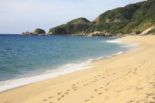 A sandy beach with gentle ocean waves, footprints leading along the shore, and green hills covered with trees in the background under a partly cloudy sky.