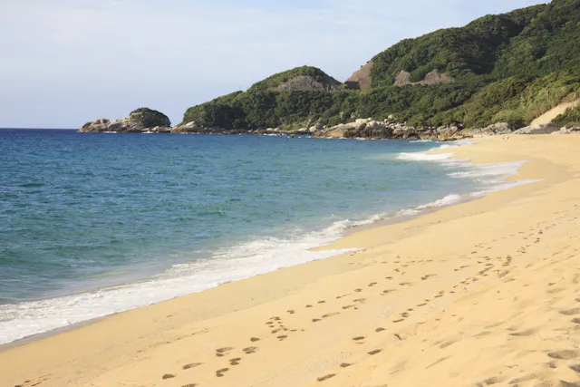 A sandy beach with gentle ocean waves, footprints leading along the shore, and green hills covered with trees in the background under a partly cloudy sky.