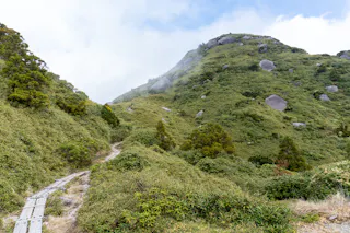 A narrow wooden trail winds through lush green shrubs and trees up a misty mountain slope, with large boulders scattered across the hillside under a partly cloudy sky.