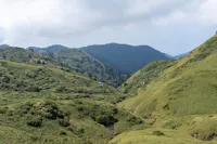 A scenic view of green, rolling hills and mountains covered with grass and shrubs under a cloudy sky.