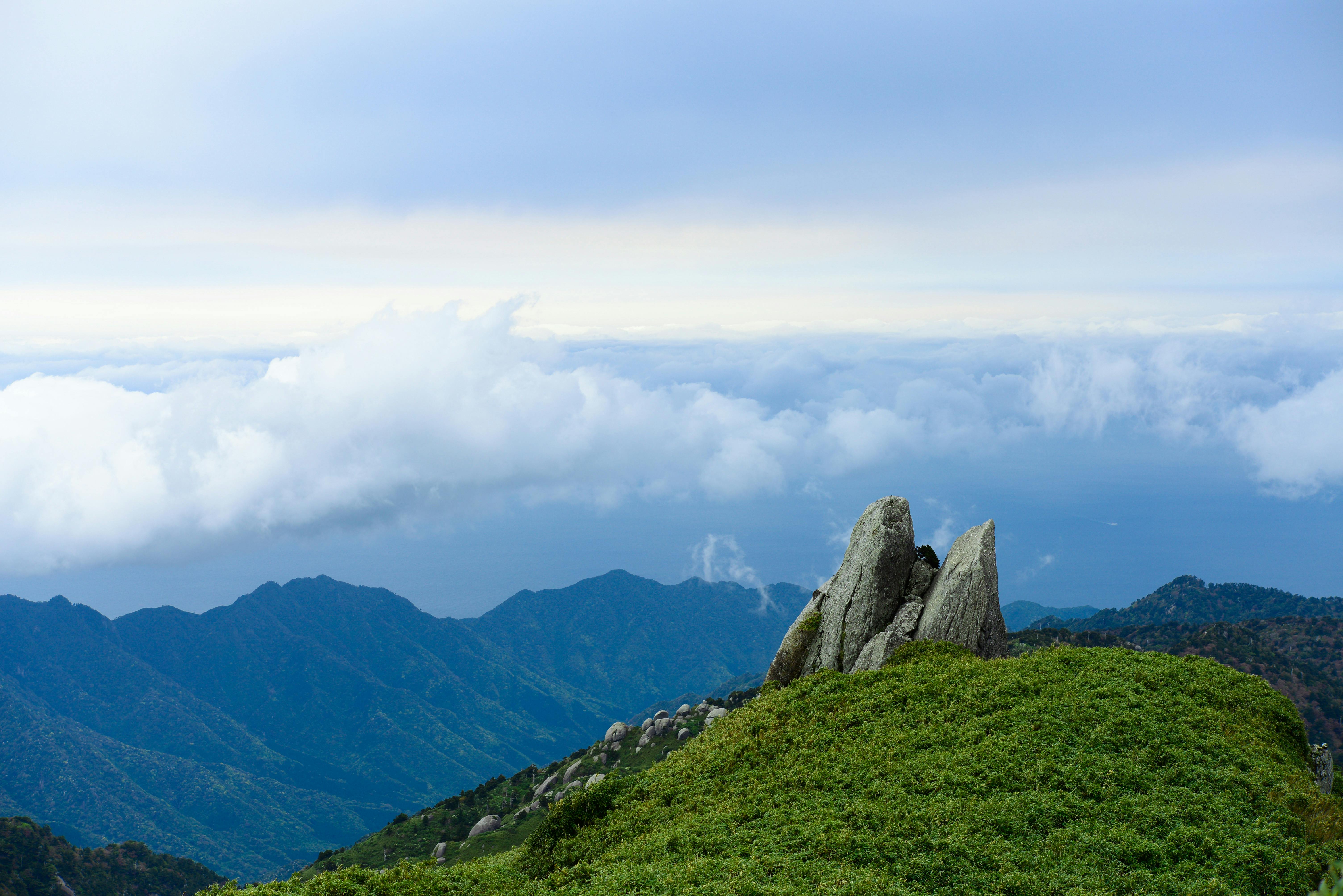 A scenic mountain landscape with green hills, rocky outcrops, and distant blue mountains under a cloudy sky. The horizon meets a layer of clouds above the mountains.