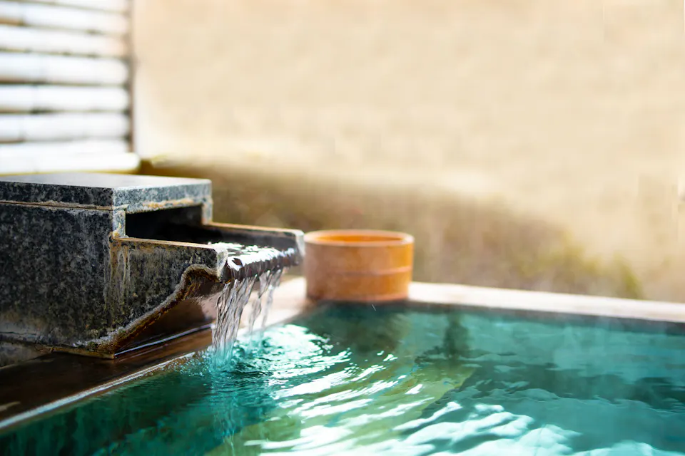A serene hot spring bath with a wooden and stone structure pouring water into a steaming, turquoise pool. The background features soft lighting and wooden panels, creating a peaceful and inviting atmosphere.
