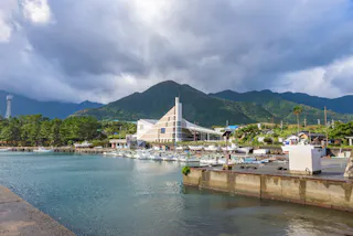 A harbor with docked boats, a modern triangular building, lush green mountains in the background, and a partly cloudy sky overhead.