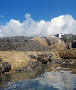 A natural rocky hot spring with clear water in the foreground, large boulders, and a small white building on the rocks under a bright blue sky with fluffy clouds.