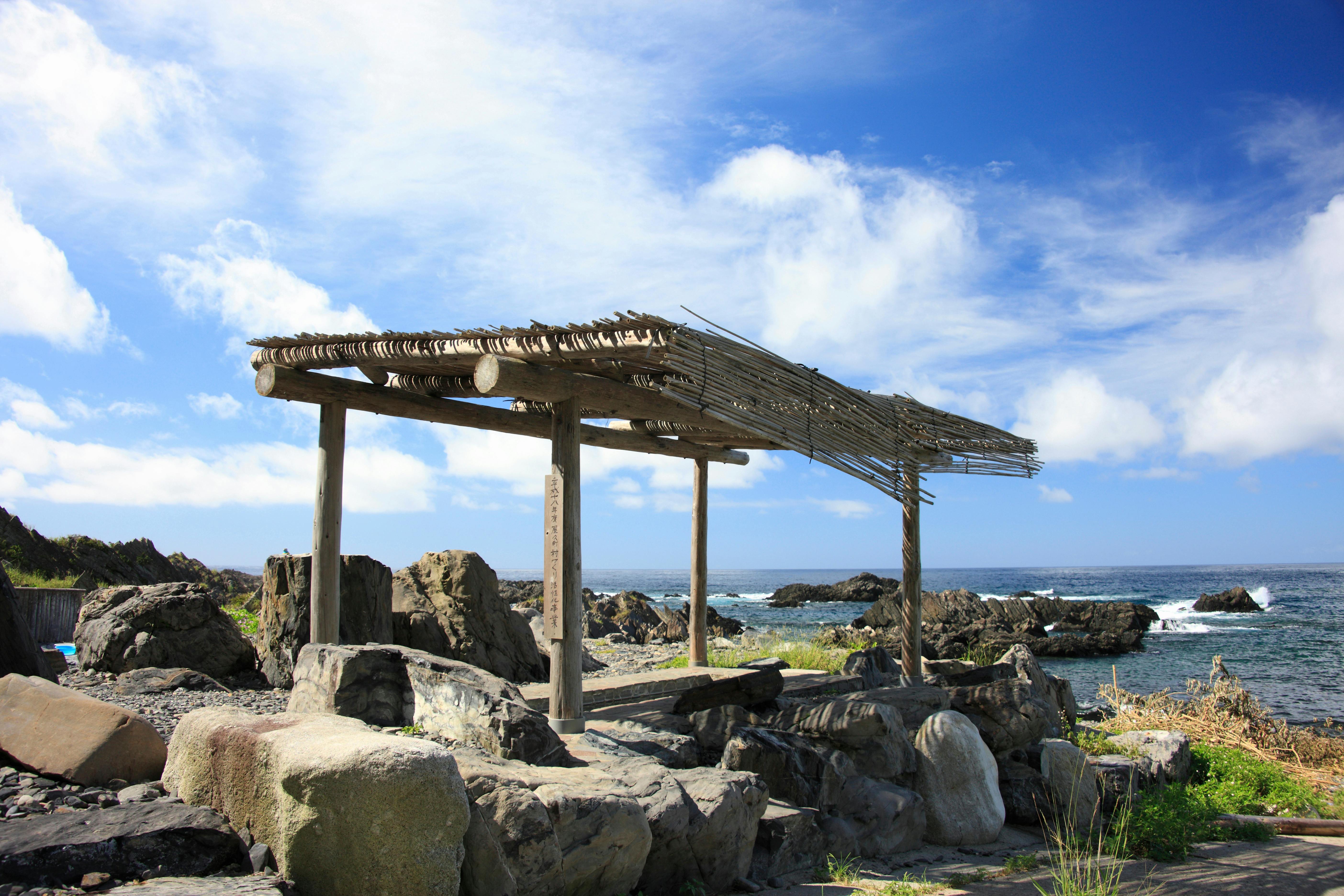 A rustic wooden shelter with a thatched roof stands among large rocks by the sea under a bright blue sky with scattered clouds. Waves crash gently against the rocky shoreline in the background.