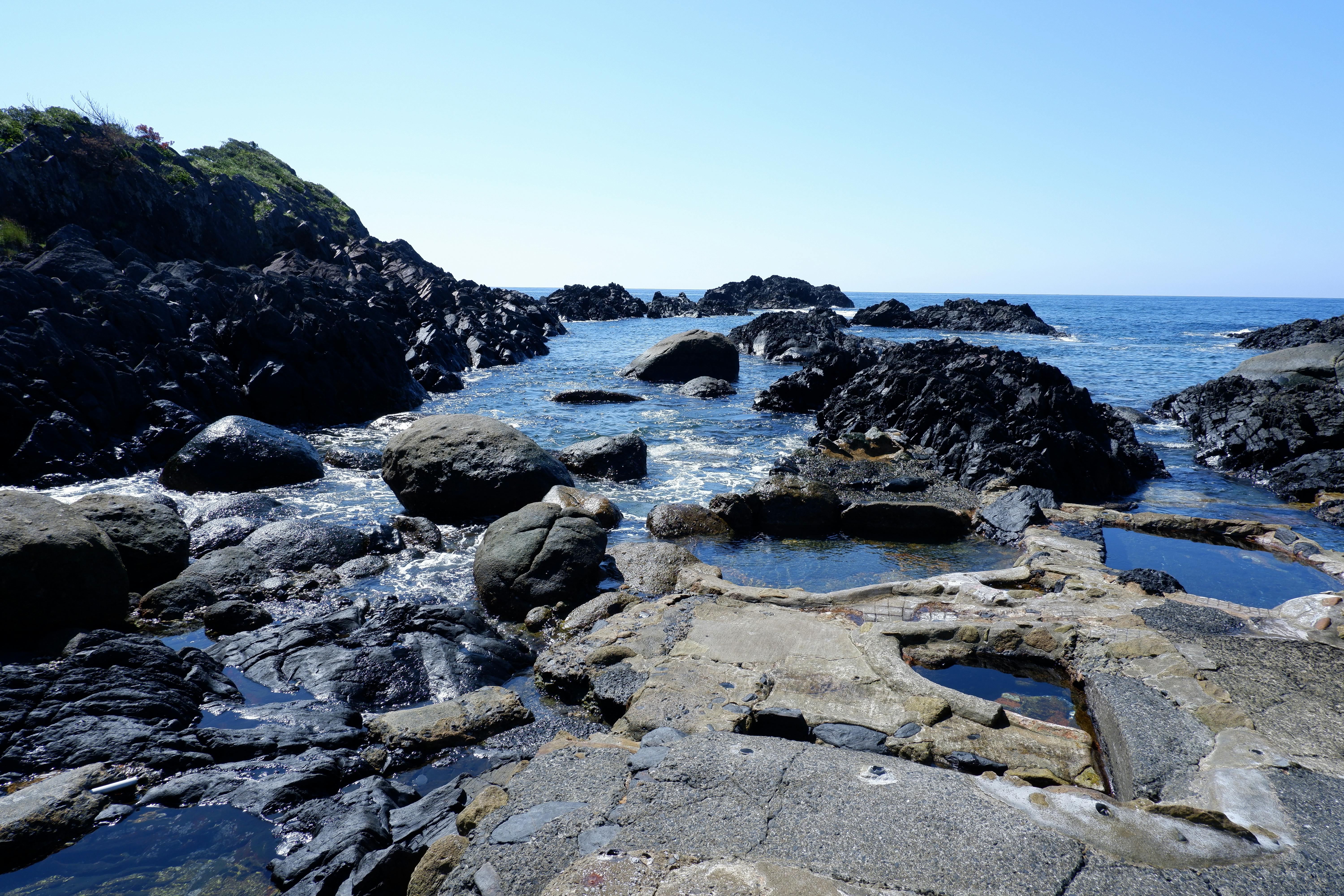 Rocky shoreline with large dark rocks and small tidal pools, leading out to a calm blue sea under a clear, bright sky. Sparse greenery grows on a hill to the left.