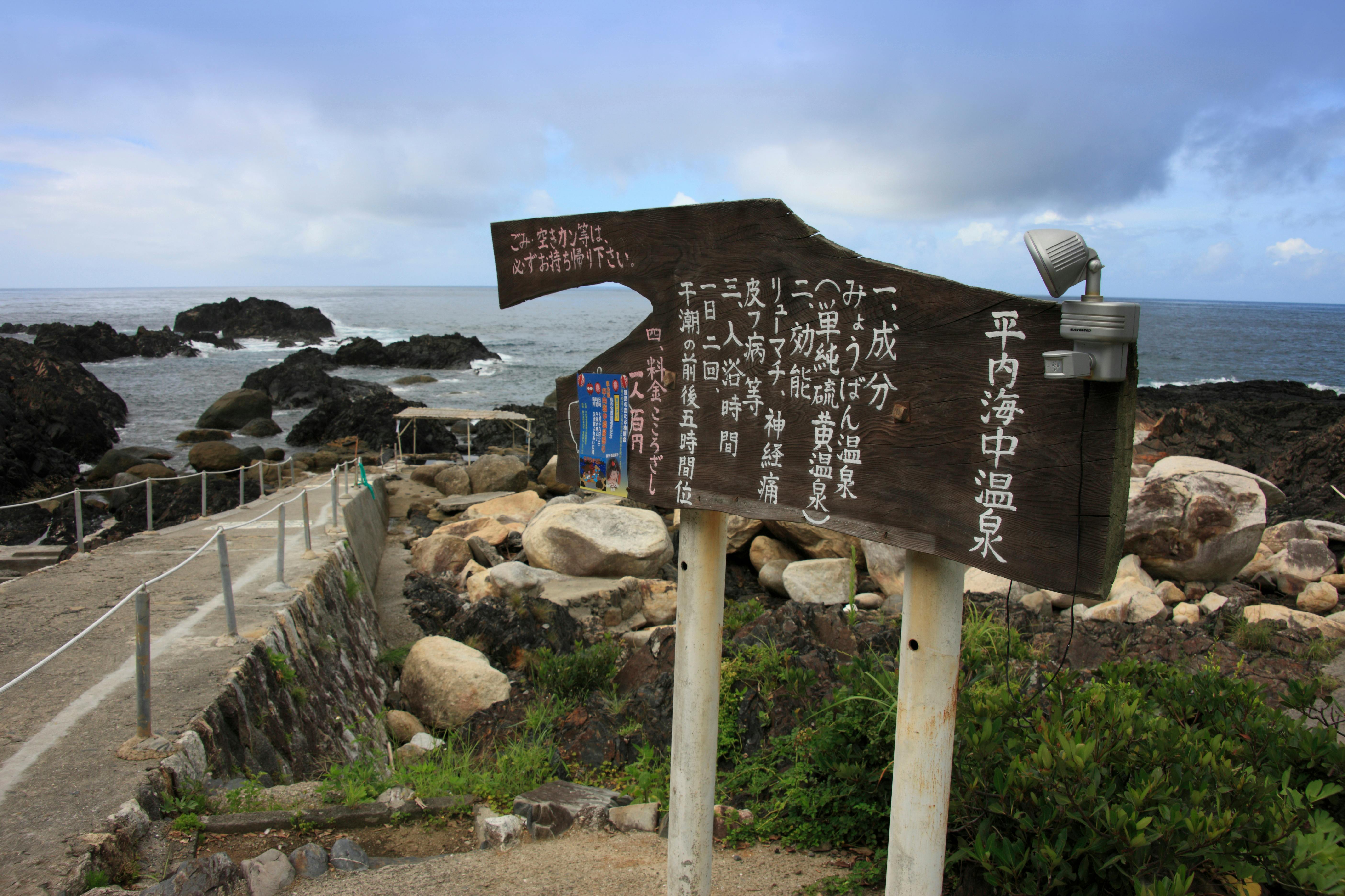 A weathered wooden sign with Japanese text stands by a rocky seaside path, leading to the ocean. Large stones, greenery, and a rope fence line the pathway under a cloudy sky.