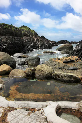 Rocky shoreline with dark jagged rocks, a natural tidal pool in the foreground, and waves crashing against the rocks under a blue sky with scattered clouds. Some greenery is visible on the distant hills.