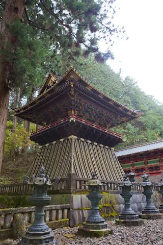 A traditional Japanese pagoda with intricate wooden carvings and gold accents stands among tall trees, surrounded by stone lanterns and moss-covered stonework in a forest setting.
