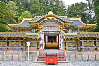 A richly decorated Japanese temple with ornate gold details, colorful carvings, and a traditional tiled roof, surrounded by lush green trees. A red offertory box stands in front of the entrance.