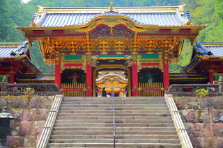 A grand, ornate Japanese temple gate with vibrant red, gold, and green details sits atop stone steps, surrounded by lush green trees in the background.