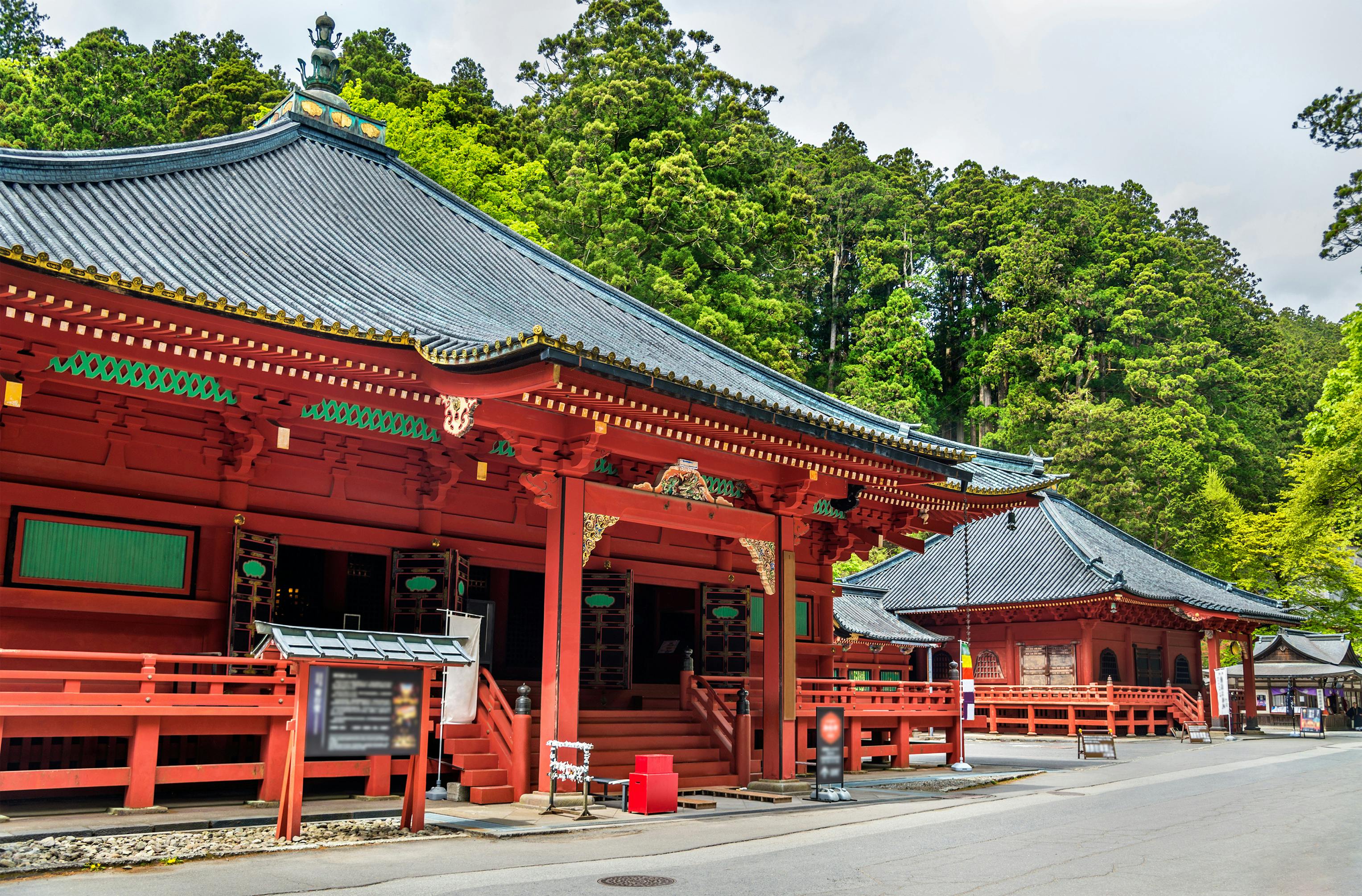 Traditional Japanese temple buildings with red wooden structures and ornate roofs, set against a backdrop of lush green trees under a partly cloudy sky. A paved path runs alongside the buildings.