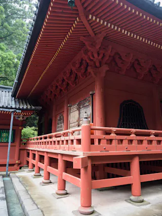 A traditional Japanese shrine with red wooden railings and ornate roof details, surrounded by green trees.