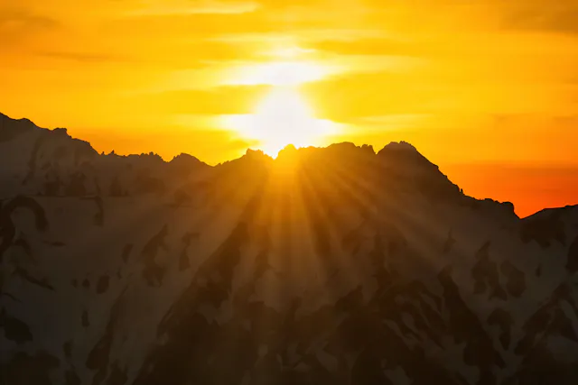 The sun sets behind snow-capped mountain peaks, casting golden light and rays across the sky and illuminating the clouds with warm shades of orange and yellow.