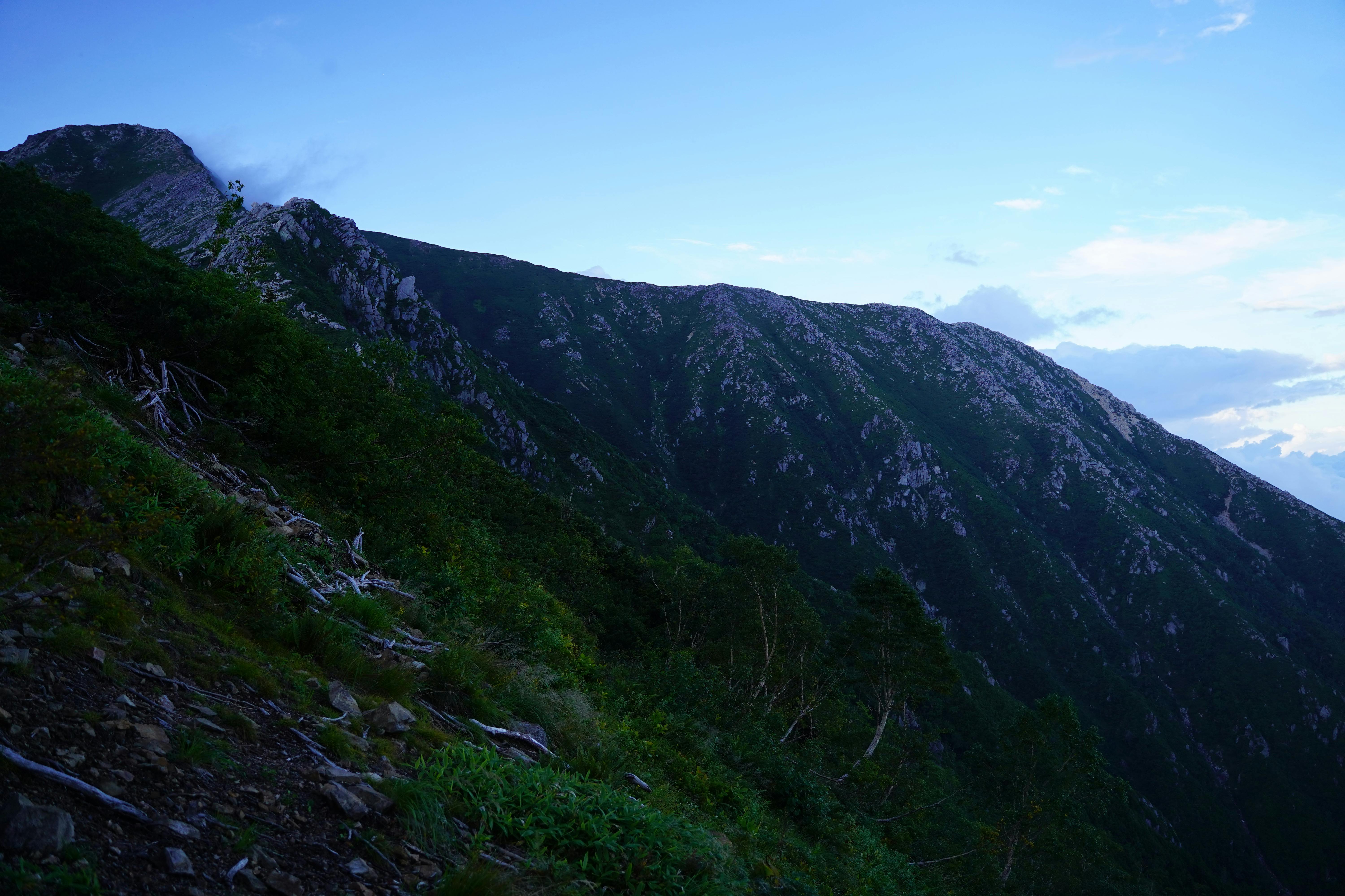 Steep, green mountain slopes under a blue sky with scattered clouds. Rocky outcrops are visible along the ridge, and dense vegetation covers the lower parts of the mountain. The scene is calm and natural.
