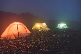 Three illuminated tents in orange, yellow, and green glow in a foggy, rocky outdoor setting at night, with mist and silhouettes of bushes in the background.