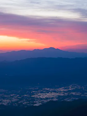 A colorful sunrise casts pink, orange, and purple hues over distant mountains and a valley below, with reflective patches of water scattered across the landscape.