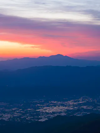 A colorful sunrise casts pink, orange, and purple hues over distant mountains and a valley below, with reflective patches of water scattered across the landscape.