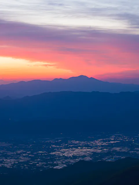 A colorful sunrise casts pink, orange, and purple hues over distant mountains and a valley below, with reflective patches of water scattered across the landscape.