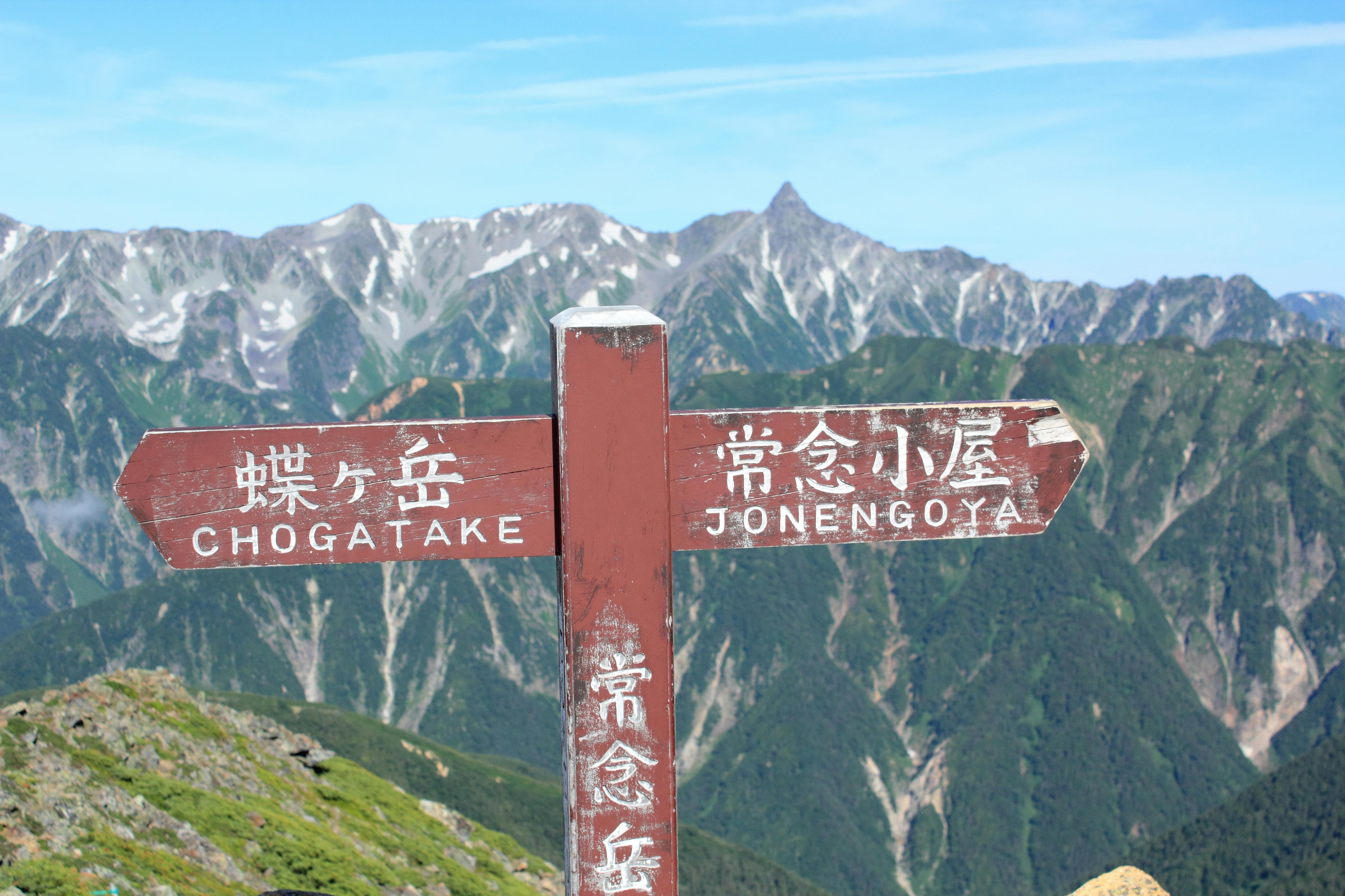 A wooden signpost with Japanese characters and English text points to “Chogatake” and “Jonengoya,” set against a backdrop of lush green mountains with rocky peaks under a clear blue sky.