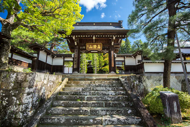 Stone steps lead to a traditional Japanese wooden gate surrounded by white walls, lush green trees, and blue sky, creating a serene temple entrance.