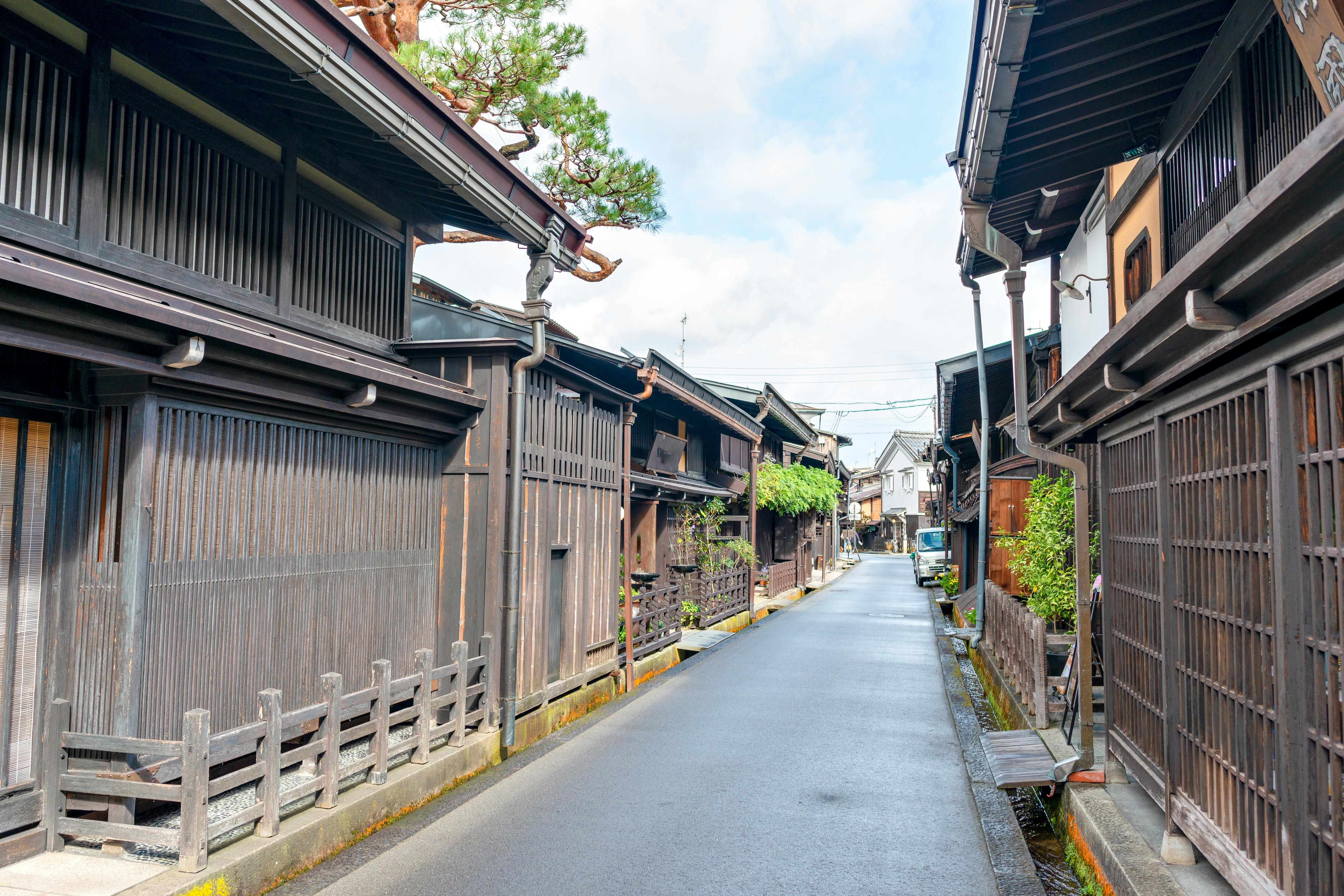 A narrow, empty street lined with traditional Japanese wooden houses, featuring dark wood facades and tiled roofs, under a partly cloudy sky. A small tree and greenery are visible along the street.