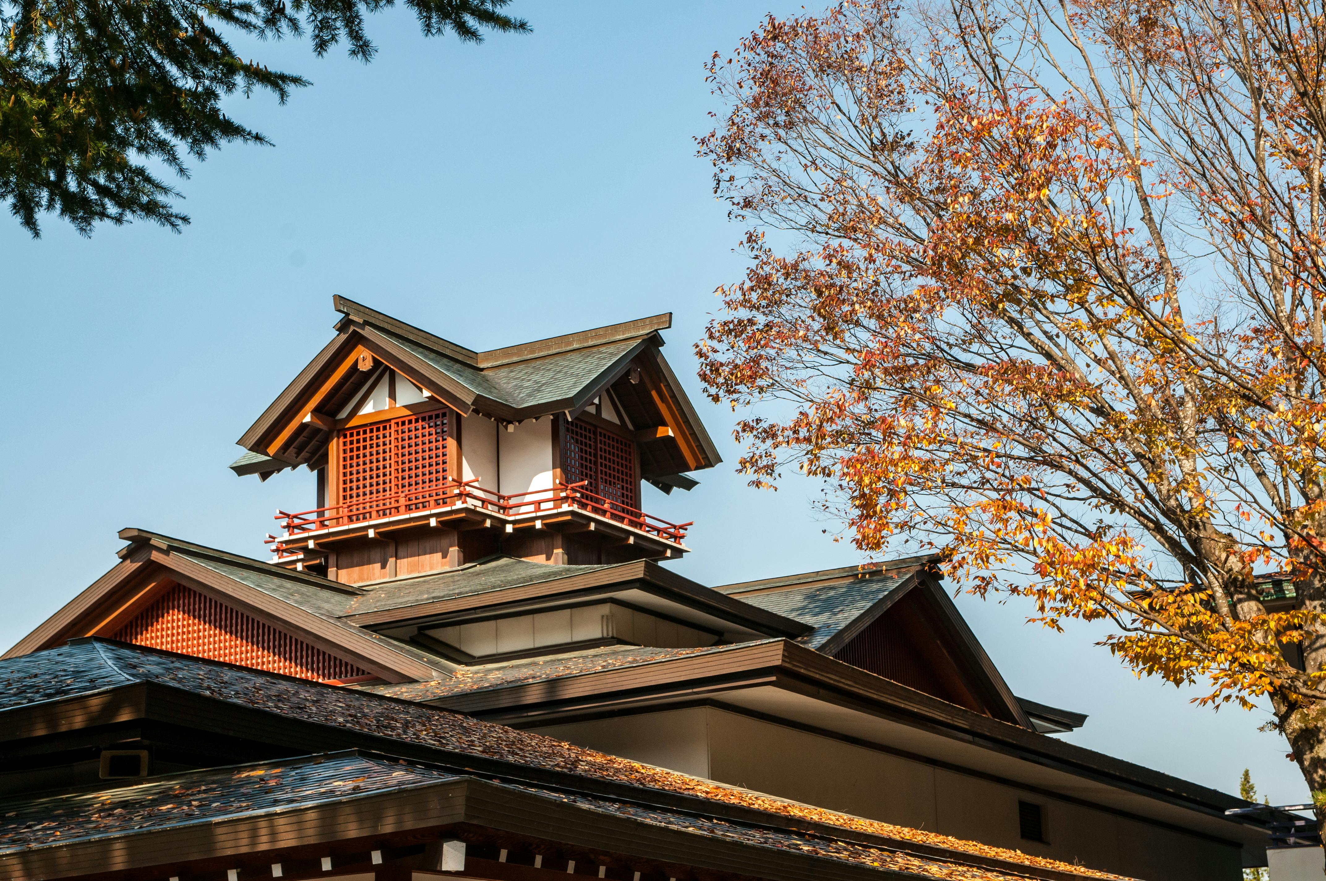 A traditional Japanese building with a prominent tower and sloped roofs, surrounded by autumn trees with orange and yellow leaves under a clear blue sky.