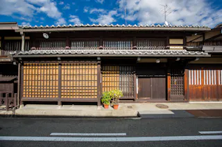 Traditional Japanese wooden house with sliding lattice doors, a tiled roof, and two potted plants outside on a sunny day under a blue sky with scattered clouds.