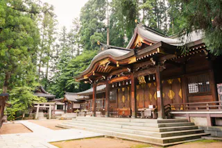 A traditional Japanese Shinto shrine with ornate wooden architecture and curved roofs, surrounded by tall green trees, sits peacefully on a stone courtyard.
