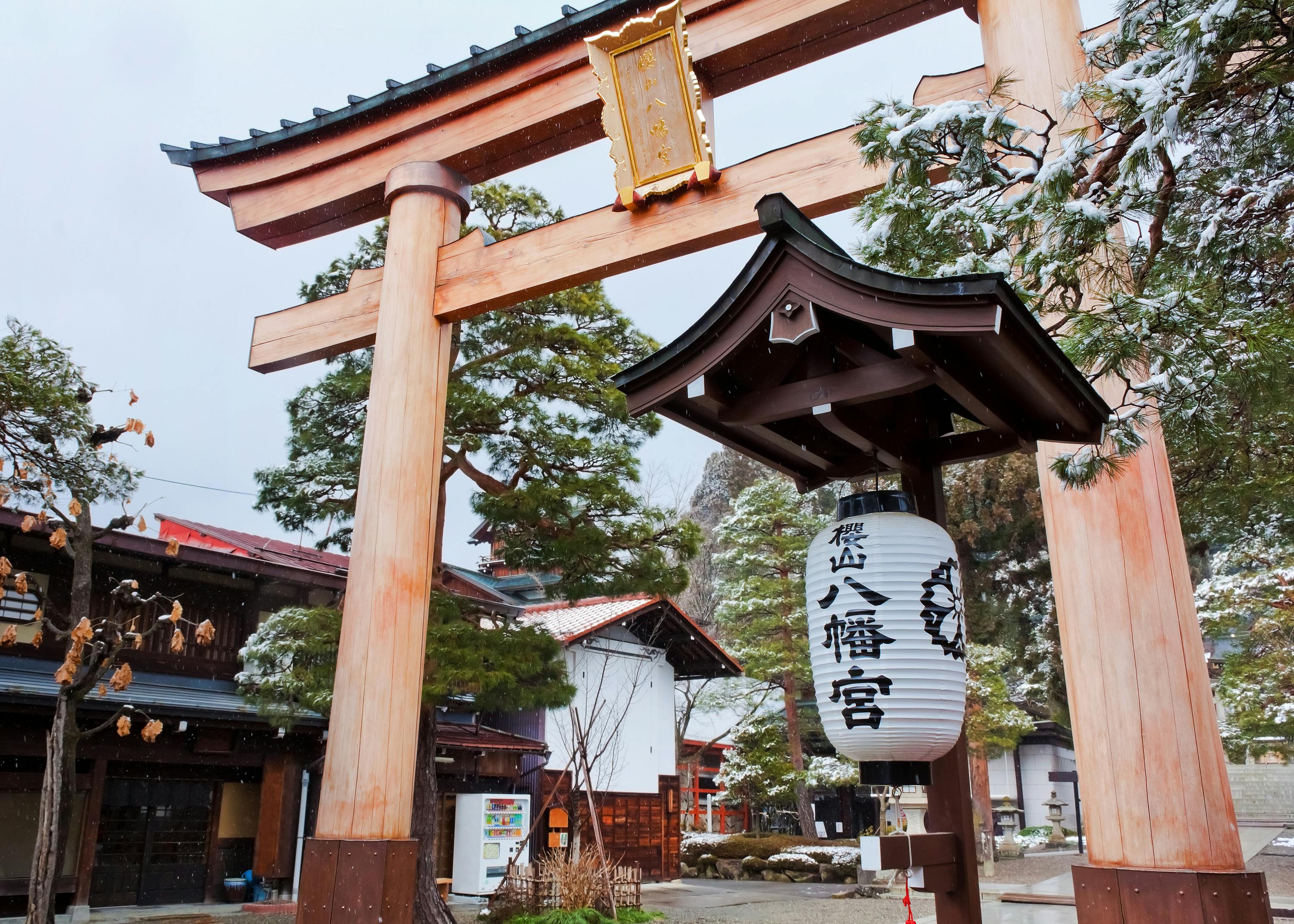 A traditional Japanese torii gate stands in a snowy landscape, with a paper lantern displaying kanji characters. Behind the gate, there are buildings and trees lightly dusted with snow. The scene conveys a serene, winter atmosphere.