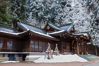 Traditional Japanese shrine with dark wooden architecture and ornate curved roofs, partially covered in snow. Snow piles and protective wooden frames are visible in front, with snow-dusted trees in the background.