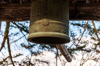 A large, ornate bronze bell with decorative carvings and inscriptions hangs outdoors from wooden beams, surrounded by trees with branches silhouetted against the sky.