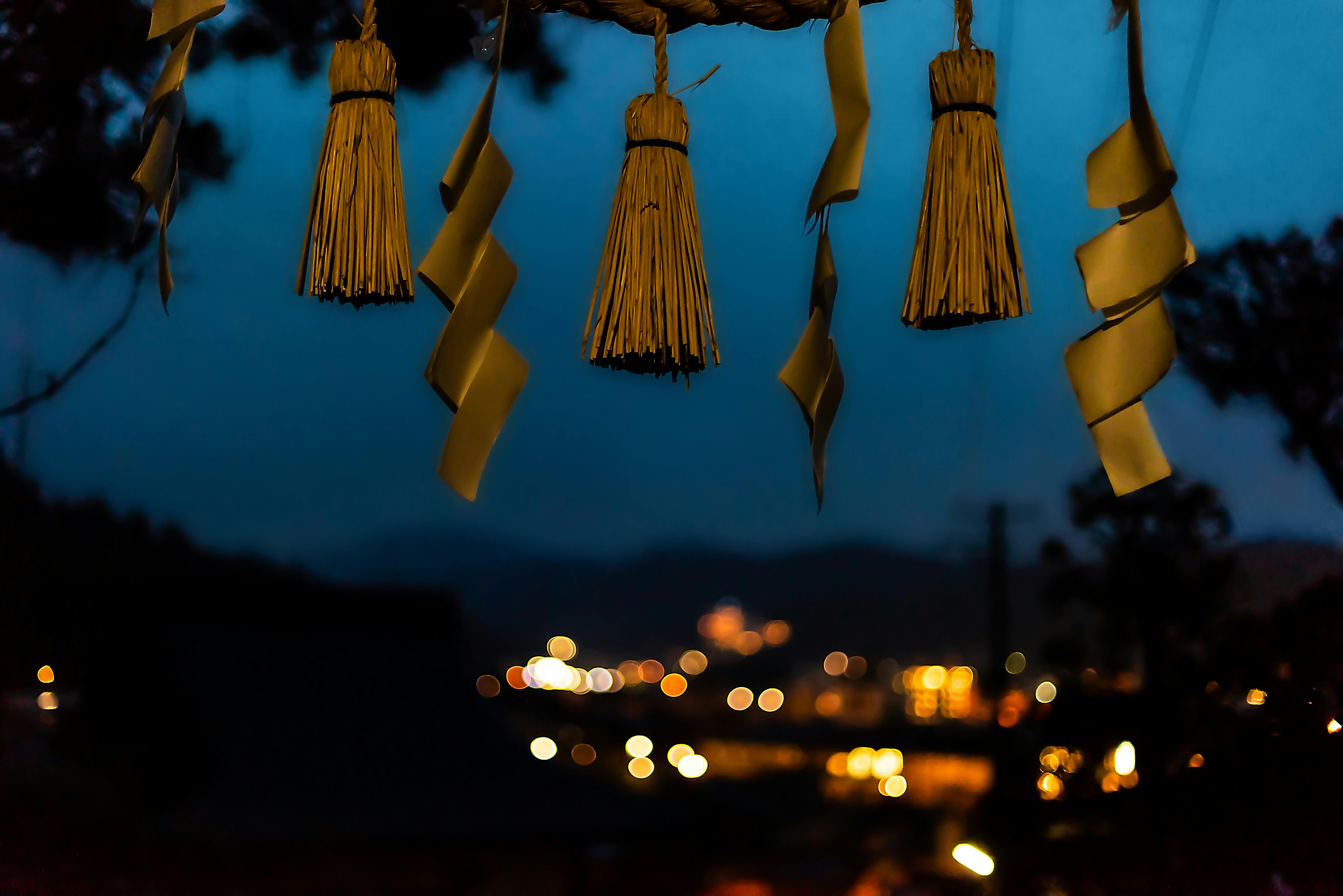 Four straw tassels and zigzag paper strips hang in the foreground, silhouetted against a dusky evening sky with blurred city lights and distant mountains in the background.