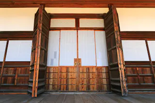 Large wooden sliding doors open to reveal shoji-style panels on a traditional Japanese building, with a wooden floor and detailed woodwork around the entrance.