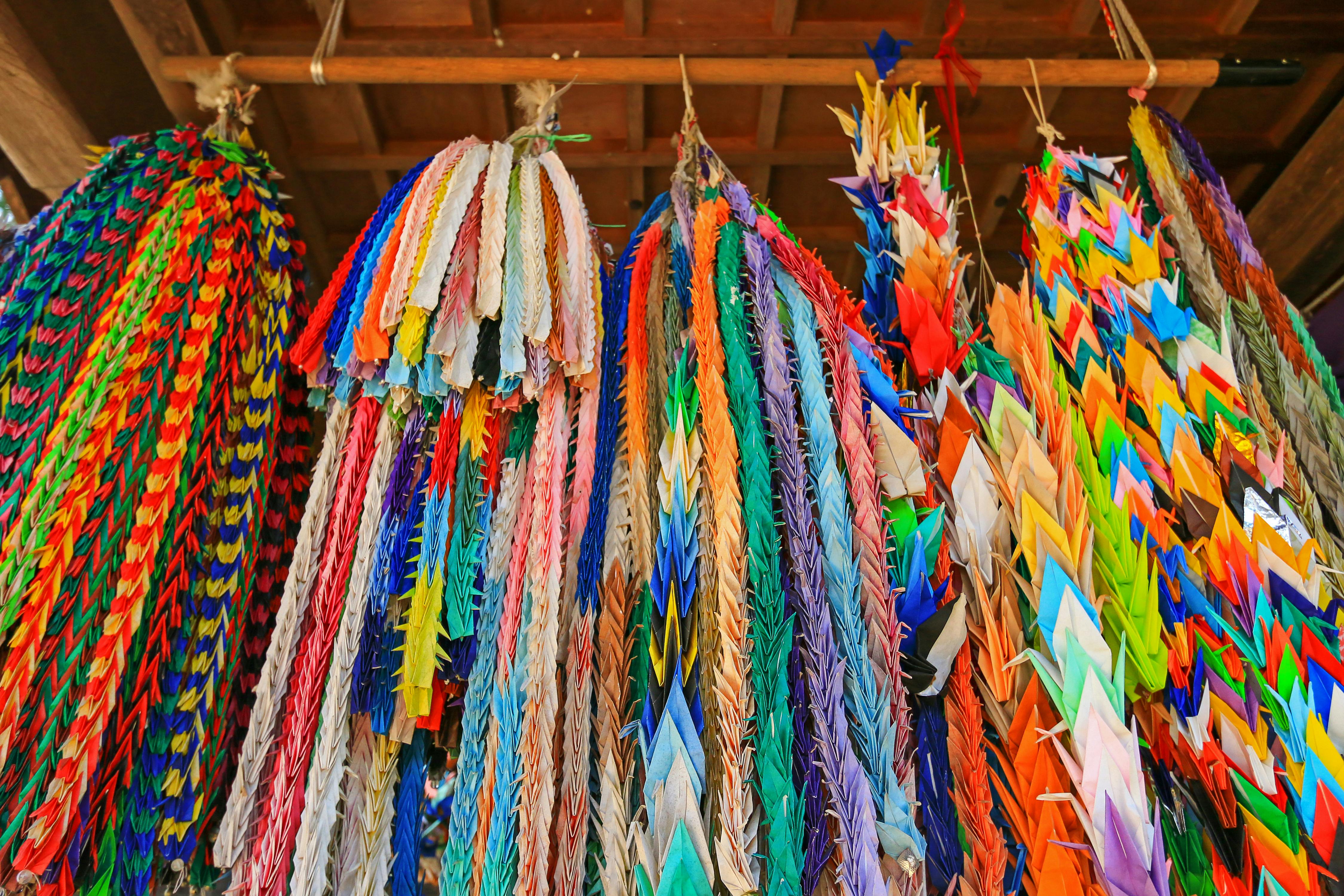 Colorful strings of paper origami cranes hang side by side, creating a vibrant display. The cranes are folded from various shades of paper and are suspended from wooden rods against a wooden ceiling.